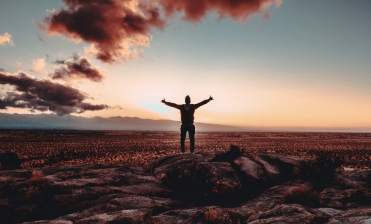 An image of a man standing on a rock with arms outspread in front of a sunset.